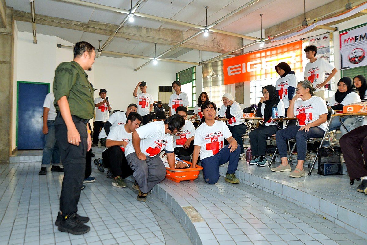 Peserta berjongkok mengelilingi manekin dalam wadah oranye saat sesi praktik pertolongan pertama, peserta lain menyaksikan dari kursi.