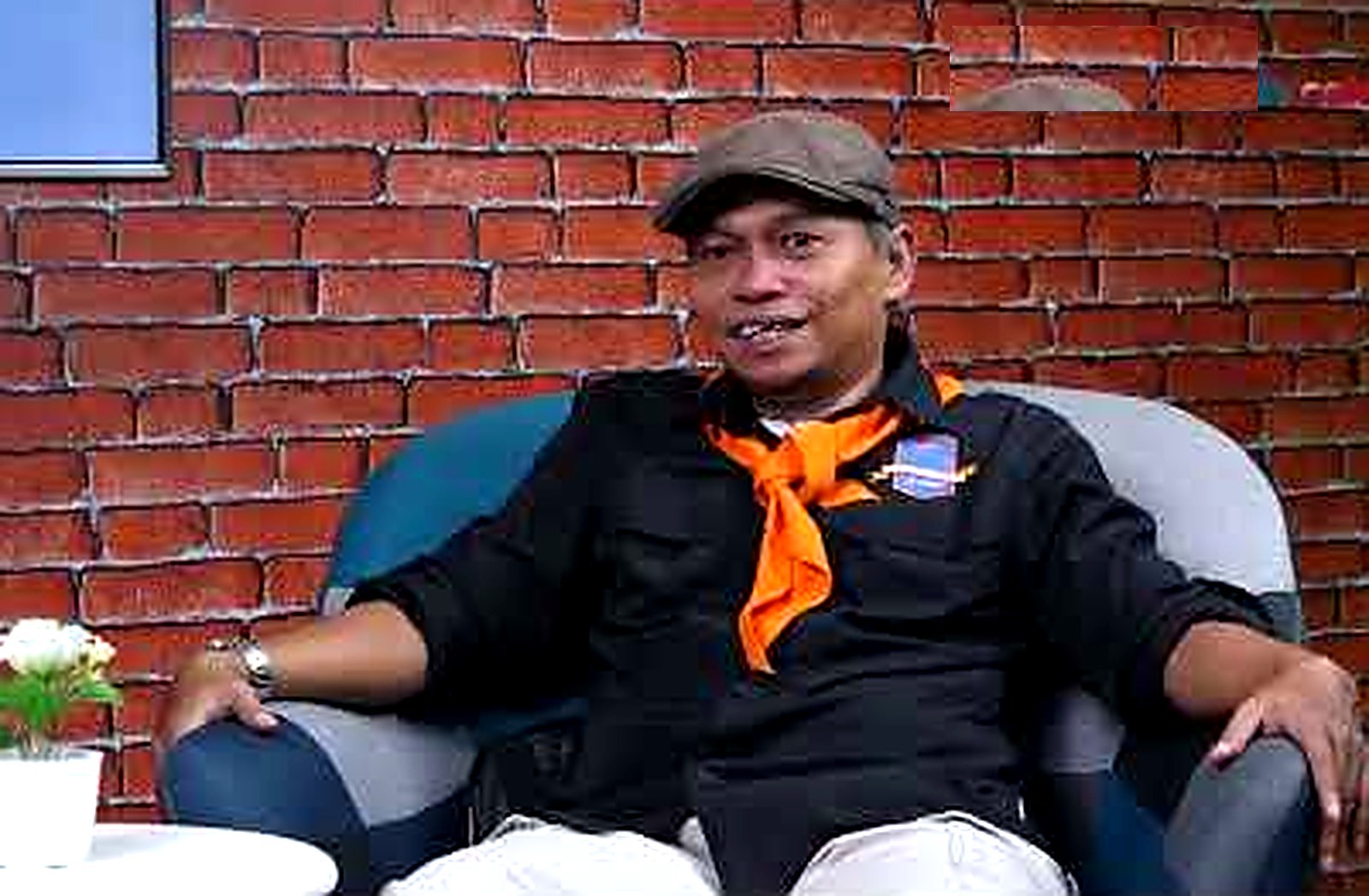 Seorang pria bertopi dan berkacu oranye duduk di kursi biru di depan dinding bata merah