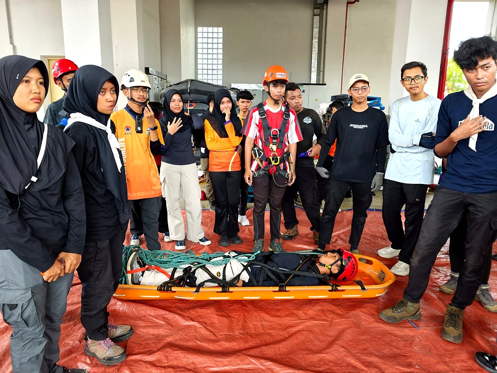 Sejumlah peserta berhelm dan berharness berdiri mengelilingi korban uji yang dibaringkan di tandu di atas terpal merah
