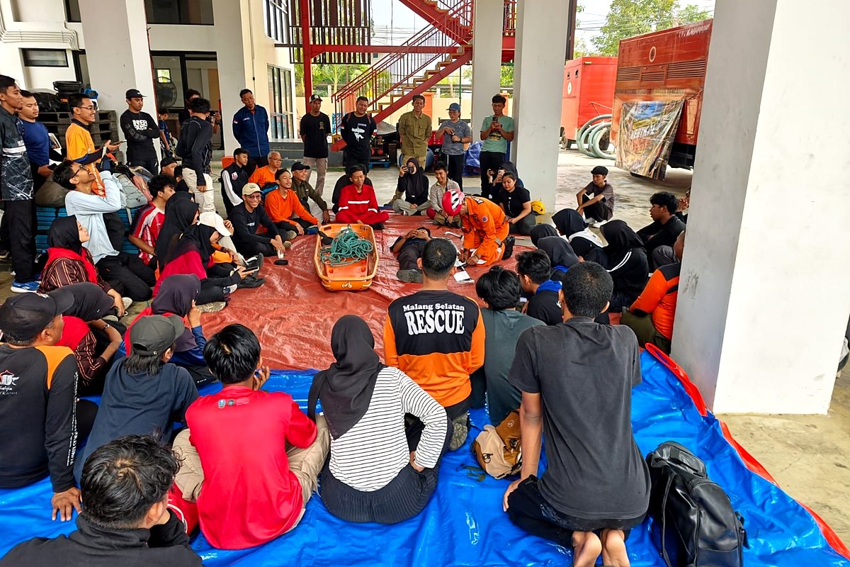 Peserta duduk melingkar di atas terpal biru dan merah dengan tandu serta tali di tengah, di bawah tangga besi gedung