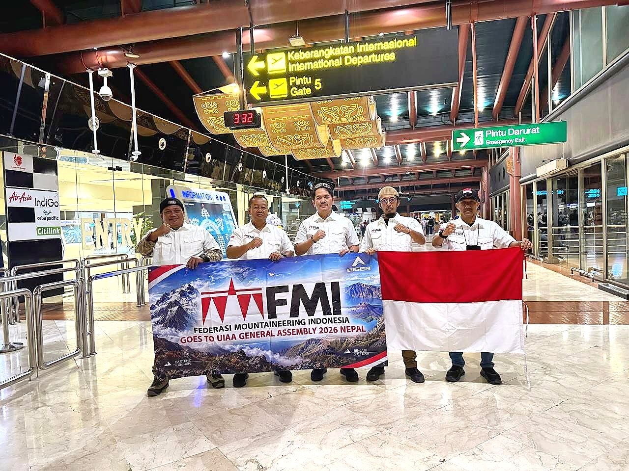 Lima pria berkemeja putih memegang spanduk FMI Goes to UIAA General Assembly 2026 Nepal dan bendera Merah Putih di area keberangkatan bandara