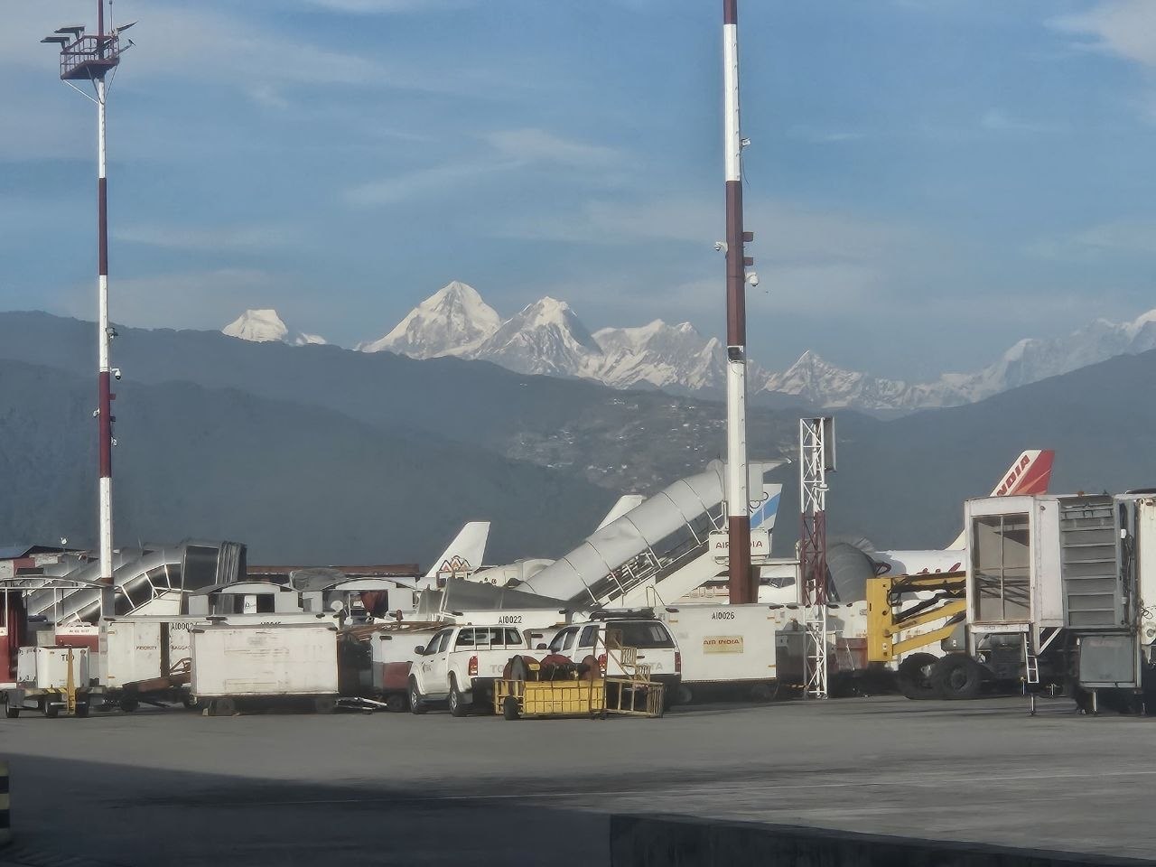 Deretan puncak Himalaya bersalju terlihat dari apron bandara, dengan peralatan darat pesawat dan tiang lampu di latar depan