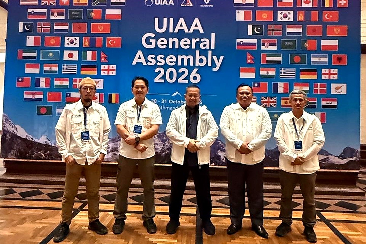 Lima anggota delegasi FMI berbaju putih berfoto di depan latar Sidang Umum UIAA 2026 bergambar bendera berbagai negara