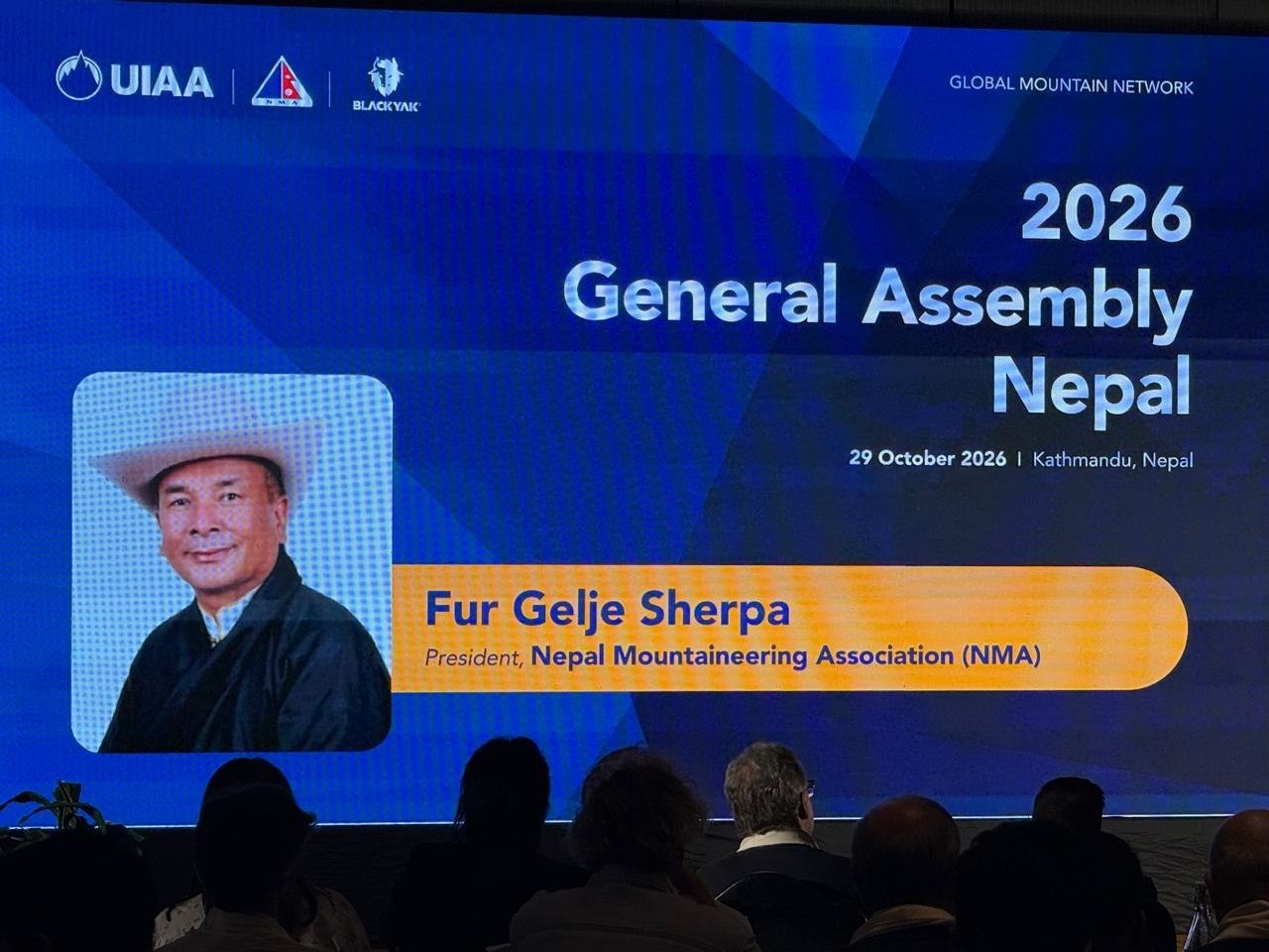 Layar panggung biru menampilkan potret dan nama Fur Gelje Sherpa, Presiden Nepal Mountaineering Association, dengan tulisan 2026 General Assembly Nepal