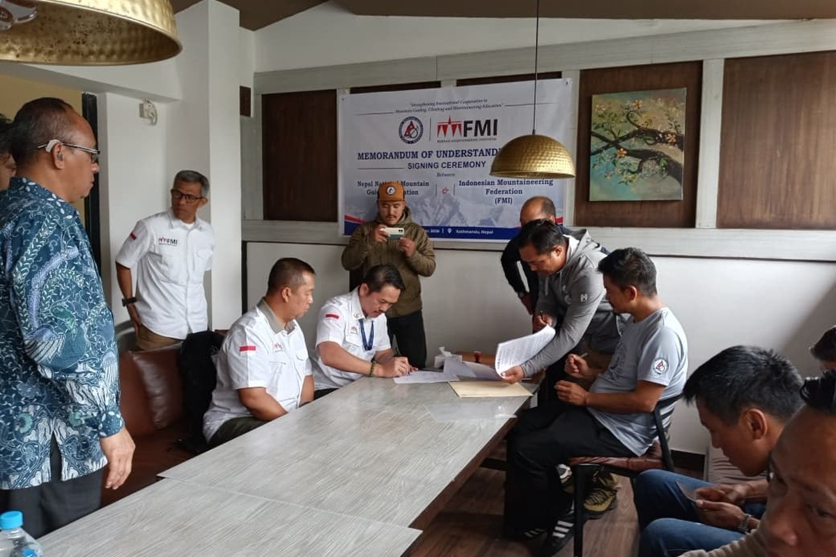 Dua orang duduk menandatangani naskah di meja, disaksikan beberapa orang berdiri di sekeliling